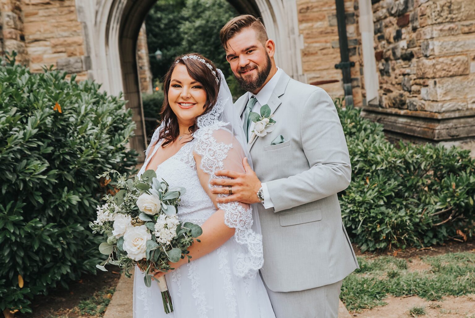 Elopement Wedding A bride and groom stand together outside a stone building on their wedding day. The bride wears a white lace wedding dress and holds a bouquet of white flowers. The groom is dressed in a light gray suit with a white shirt and matching boutonnière. Both are smiling at the camera. Elopements Inc